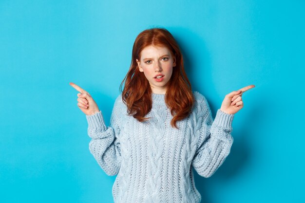 Rousse confuse en pull pointant les doigts sur le côté, regardant la caméra dubitative, debout sur fond bleu. Espace de copie