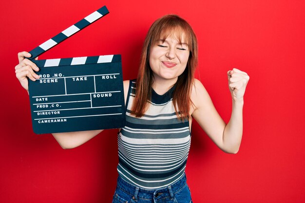 Rousse jeune femme tenant un film vidéo à clin criant fier, célébrant la victoire et le succès très excité avec le bras levé