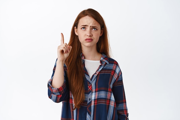 Une rousse sceptique pointe du doigt quelque chose de boiteux, grimaçant et fronçant les sourcils contrarié, pointant et regardant avec regret, chose injuste, debout sur fond blanc