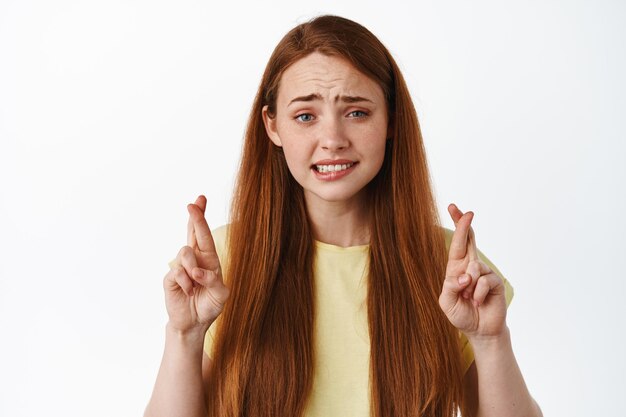 Une rousse stressée et nerveuse croise les doigts et a l'air inquiète, prie, fait un vœu, attend anxieusement des résultats importants, debout sur fond blanc.