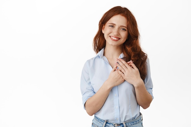 Une rousse touchée et ravie dit merci, regarde quelque chose de flatteur et de gentil, se tenant la main sur le cœur, exprime sa gratitude et ses remerciements, sourit sincèrement, debout sur fond blanc