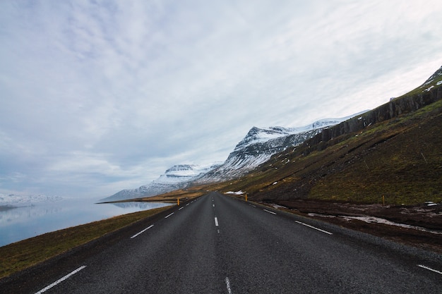 Route entourée par la rivière et les collines couvertes de neige et d'herbe sous un ciel nuageux en Islande