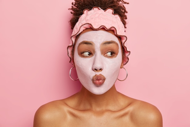 Photo gratuite routine de soin. la belle femme afro-américaine garde les lèvres arrondies, applique un masque nourrissant sur le visage, réduit les risques d'acné, obtient peu de plaisir dans la vie grâce aux traitements de beauté, améliore la peau