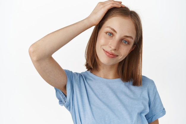 Routine de soins de la peau et beauté. Heureuse jeune fille blonde touche sa coiffure, souriant et regardant tendre la caméra, joli visage avec éclat naturel et sans maquillage, fond blanc