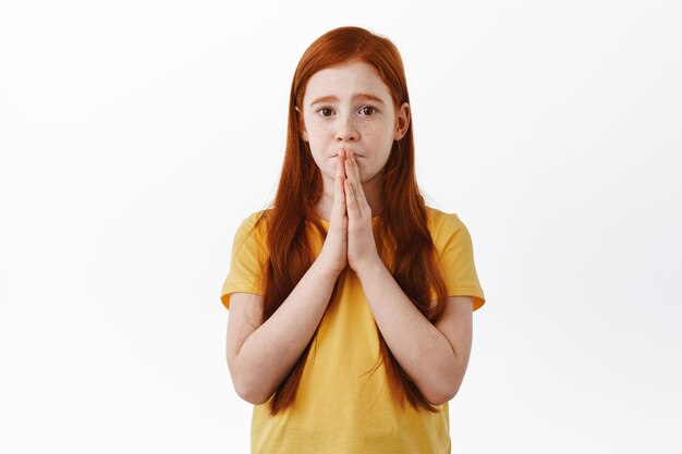 S'il te plaît. Petite fille mignonne faisant un visage timide et nécessiteux et demandant quelque chose, suppliant d'acheter un jouet, suppliant les mains de prier, regardant avec des yeux tristes la caméra, fond blanc