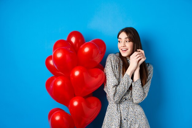 La Saint-Valentin. Belle fille romantique rêvant de date, debout près de jolis ballons coeur et souriant, fond bleu.