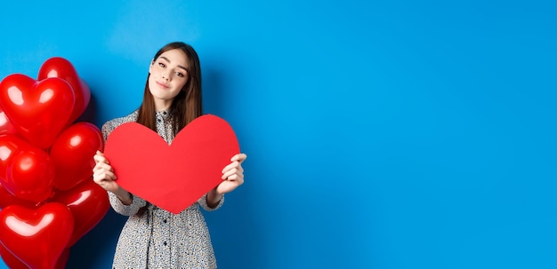 Saint valentin fille romantique en robe montrant une grande découpe de coeur rouge rêvant d'amour debout près de ho