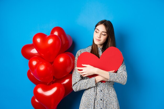 La Saint-Valentin. Jolie femme romantique en robe étreignant la grande découpe de coeur rouge et à la recherche de rêve, pensant à l'amour, debout sur fond bleu.