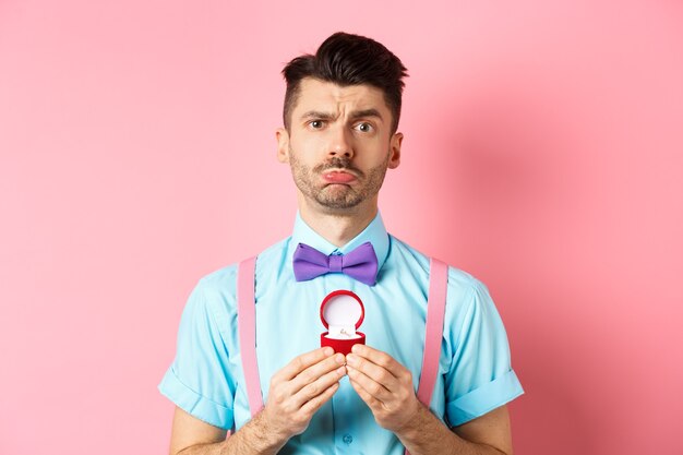 La Saint-Valentin. Triste petit ami rejeté, montrant une bague de fiançailles et boudant bouleversé, elle a dit non, debout sur fond rose.