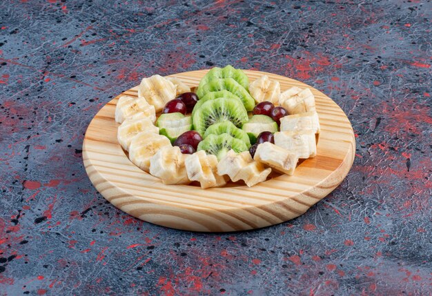 Salade de fruits avec banane tranchée, kiwis et baies dans une assiette en bois.