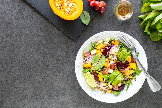 Salade de légumes frais avec betterave, roquette, oignon rouge, oseille, pois chiches, citrouille et raisins dans une assiette blanche sur un tableau noir. Vue de dessus