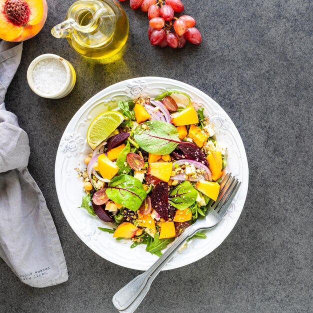 Salade de légumes frais avec betteraves, roquette, oignon rouge, oseille, pois chiches, pêche et raisins dans une assiette blanche sur fond noir. Vue de dessus