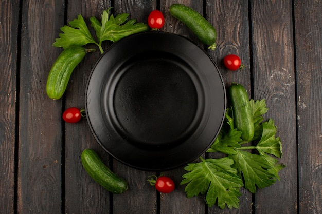 Photo gratuite salade de légumes tels que les concombres et les tomates cerises rouges avec des feuilles vertes autour d'une plaque noire sur un sol brun rustique en bois