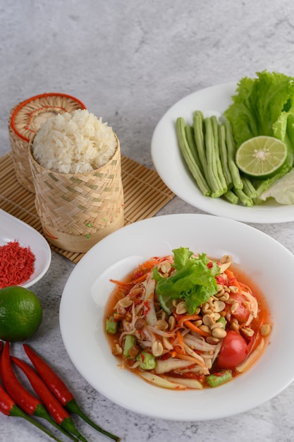 Salade de papaye thaïlandaise dans une assiette blanche avec du riz gluant dans un panier en osier de bambou et des crevettes séchées