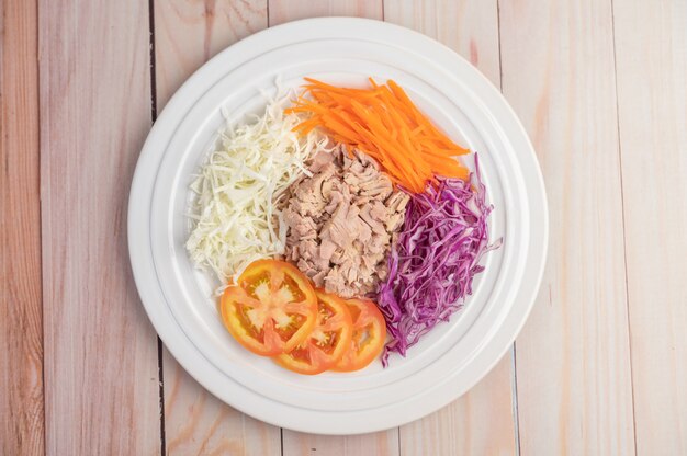 Salade de thon aux carottes, tomates, chou sur une plaque blanche sur un plancher en bois.