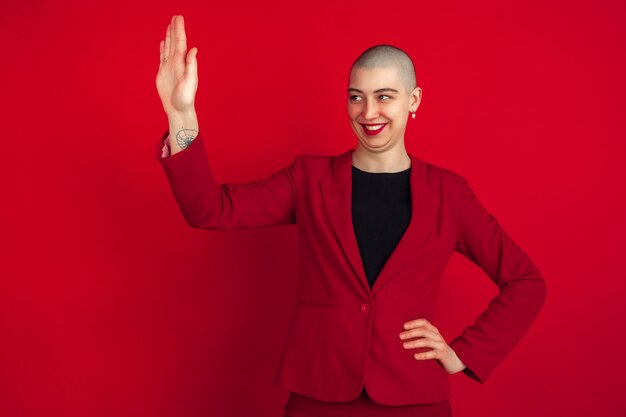 Saluer quelqu'un. Portrait de jeune femme chauve caucasienne isolée sur mur rouge. Beau modèle féminin en veste. Émotions humaines, expression faciale, ventes, concept publicitaire. Bizarre.