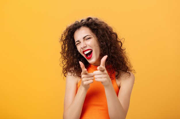 Salut quoi de neuf. Sympathique et extravertie séduisante femme aux cheveux bouclés avec du rouge à lèvres rouge pointant vers la caméra avec des pistolets à doigt comme si elle saluait ou faisait allusion à un ami faisant un clin d'œil à la caméra sur fond orange.