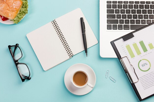 Sandwich, tasse à café, lunettes, bloc-notes à spirale, stylo, ordinateur portable et bloc-notes avec plan budgétaire sur le bureau bleu