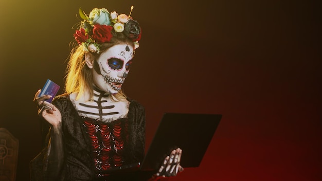 Photo gratuite santa muerte faisant des achats en ligne sur un ordinateur portable, payant par carte de crédit en studio. faire une transaction de paiement électronique sur pc, ressemblant à la sainte déesse de la mort sur la tradition d'halloween. prise de vue à main levée.