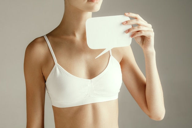 Photo gratuite santé de la femme. modèle féminin tenant une carte vide près de la poitrine. jeune fille adulte avec du papier pour signe ou symbole isolé sur fond gris studio. découpez une partie du corps. problème médical et solution.