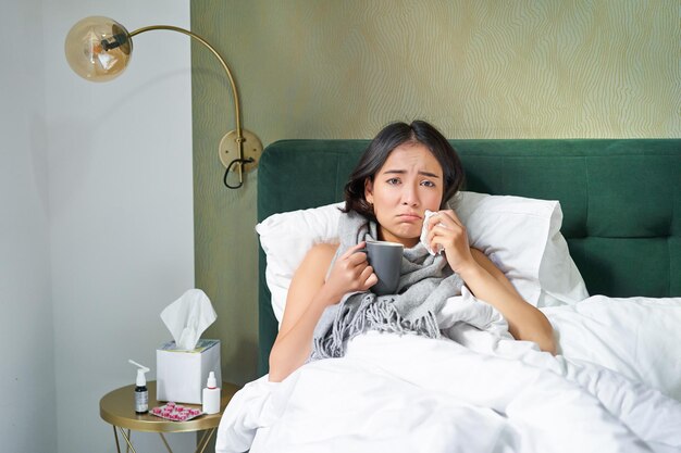Santé et personnes fille asiatique allongée dans son lit se sentant malade éternuant et buvant du thé chaud attrapant un rhume s