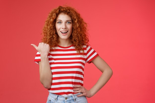 Sassy beau émotif heureux souriant rousse européenne femme bouclée coiffure pointant le pouce gauche sourire assertif effronté tenir la taille de la main dirigeant l'offre publicitaire promo fond rouge.