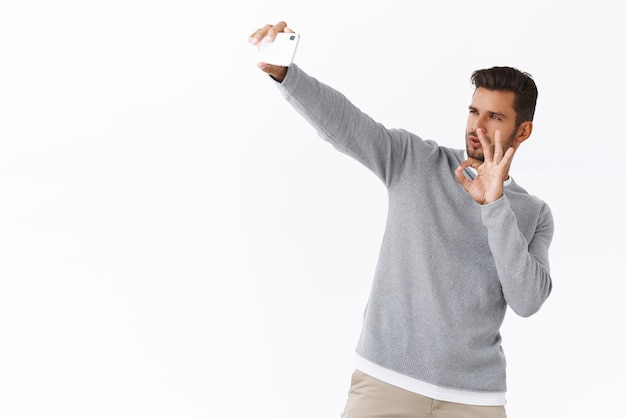 Sassy beau influenceur internet beau modèle masculin prenant selfie sur smartphone avec la main tendue faisant un geste d'approbation correct enregistrer vlog et recommander le fond blanc du produit