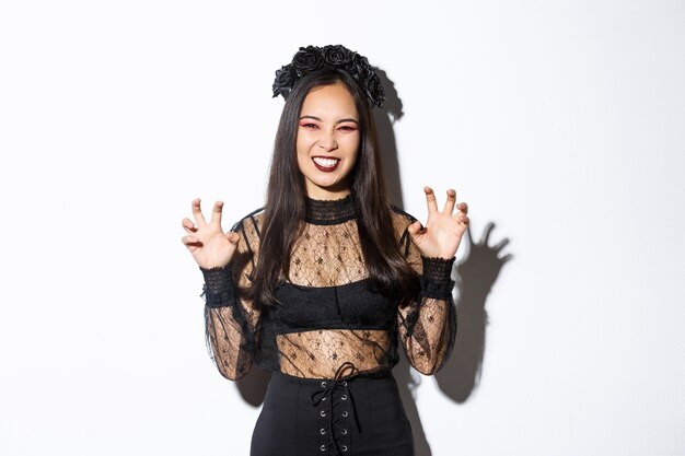 Sassy belle femme asiatique en robe de dentelle noire et guirlande essayant de vous effrayer, levant les mains, agissant comme une sorcière maléfique lors d'un événement d'halloween, debout sur fond blanc.