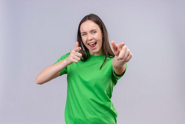 Satisfait de belle jeune fille portant un t-shirt vert sorti et heureux de pointer avec les doigts vers la caméra souriant joyeusement debout sur fond blanc isolé