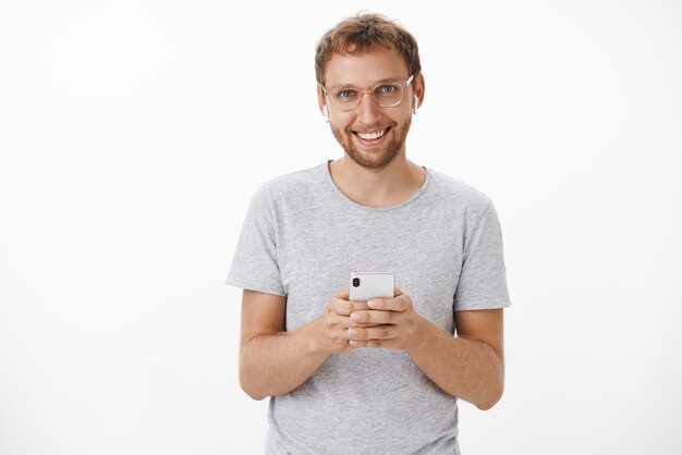 satisfait client masculin heureux avec des poils dans des lunettes et un t-shirt gris tenant un tout nouveau smartphone portant des écouteurs sans fil ravi de la bonne qualité du son