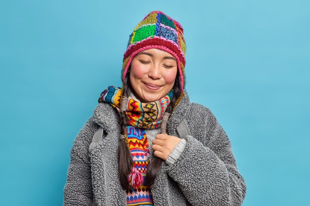 Satisfait et heureux, une femme asiatique ferme les yeux et sourit porte agréablement un bonnet tricoté et une écharpe manteau chaud pour l'hiver froid a deux tresses rappelle quelque chose d'agréable isolé sur un mur bleu