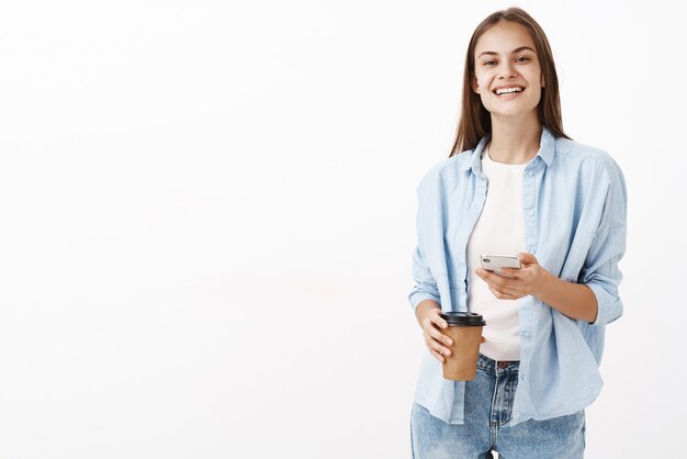 Satisfait heureux jolie femme caucasienne en chemisier bleu sur t-shirt tenant une tasse de café et téléphone portable souriant avec délice heureux d'avoir une pause tout en travaillant au bureau