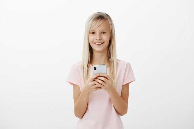 Satisfait de joyeuse fille blonde mignonne en t-shirt rose, tenant un smartphone cher et souriant