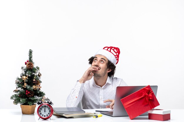 Satisfait positif jeune homme d'affaires avec drôle de chapeau de père Noël regardant au-dessus de penser et de célébrer Noël au bureau sur fond blanc