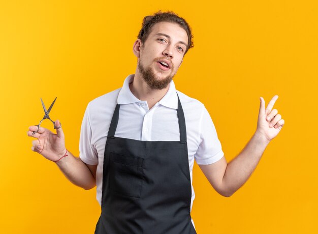 Satisfait de la tête inclinable du jeune coiffeur masculin en uniforme tenant des ciseaux sur le côté isolé sur un mur jaune avec espace pour copie