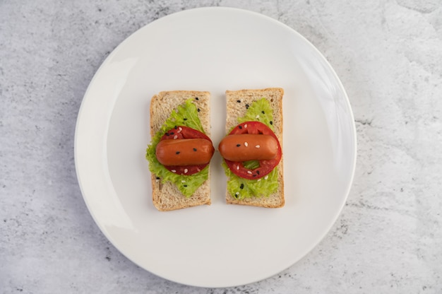 Saucisse aux tomates, salade et deux ensembles de pain sur une plaque blanche.