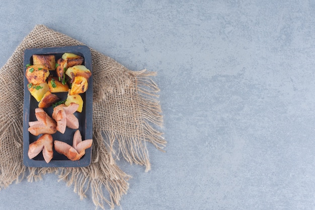 Saucisse frite aux pommes de terre sur une plaque en bois noire.