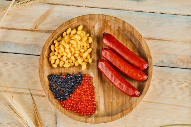 Saucisses, graines de maïs bouillies et caviar sur une assiette en bois.
