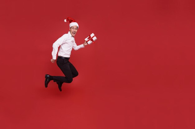 Sautant jeune homme d'affaires asiatique portant le bonnet de Noel et tenant une boîte-cadeau