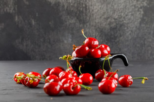 Savoureuses cerises dans une cuillère noire sur une table grungy et grise, vue latérale.