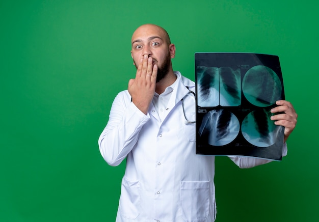 Photo gratuite scared young male doctor wearing medical robe et stéthoscope holding x-ray et bouche couverte avec main isolé sur fond vert