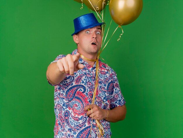 Scared young party guy portant un chapeau bleu tenant des ballons attachés autour du cou vous montrant le geste isolé sur vert