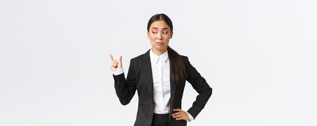 Sceptique et maladroite jeune femme d'affaires asiatique vendeuse en costume noir façonnant quelque chose de petit et ayant l'air déçue par la taille grimaçant sans amusement sur une petite chose minuscule fond blanc