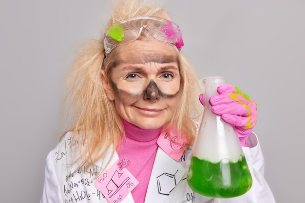 Un scientifique au travail mène une expérience en laboratoire tient de la verrerie avec un liquide vert a le visage sale après qu'une expérience chimique porte un manteau médical et des gants isolés sur fond gris