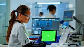 Photo gratuite scientifique de recherche médicale travaillant sur tablette avec modèle de maquette d'écran vert dans un laboratoire de sciences appliquées. ingénieurs menant des expériences en arrière-plan, examinant l'évolution des vaccins à l'aide de la haute technologie