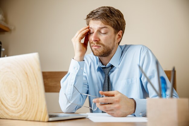 Se sentir malade et fatigué. Frustré triste malheureux jeune homme malade massant sa tête alors qu'il était assis à son lieu de travail au bureau. La grippe saisonnière, la grippe pandémique, le concept de prévention des maladies