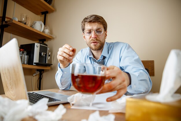 Se sentir malade et fatigué. L'homme avec une tasse de thé chaud travaillant au bureau, homme d'affaires attrapé froid, grippe saisonnière. Grippe pandémique, prévention des maladies, climatisation dans le bureau causent des maladies