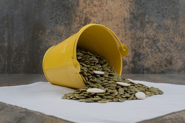 Seau jaune plein de graines de citrouille pelées sur une nappe blanche.