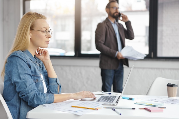 Une secrétaire blonde confiante utilise un ordinateur portable pendant que le patron a une conversation téléphonique