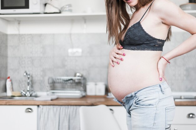 Section médiane de la femme enceinte debout dans la cuisine en touchant son ventre et le dos